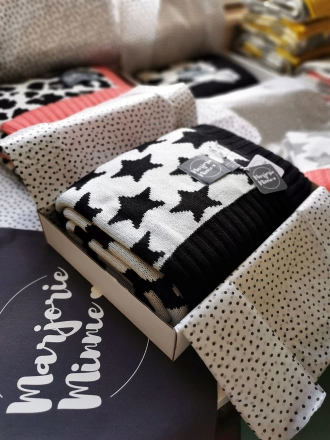 A neatly folded monochrome star blanket is shown packed inside a box and wrapped with tissue paper. The box is sitting on a table surrounded by other gift wrapped products. Beneath the box is a grey Marjorie Minnie branded gift bag.