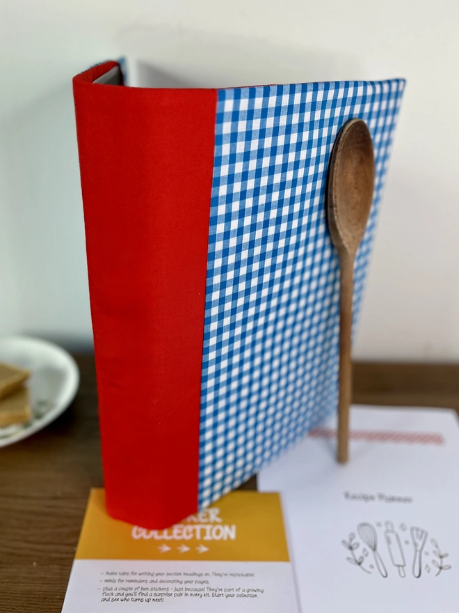 Blue Gingham Recipe Binder with plain red spine (non-embroidered) standing on wooden bench with wooden spoon propped against it & recipe template pages and 'Sticker Collection' envelope lying beside it. Home-baked squares on plate in background.