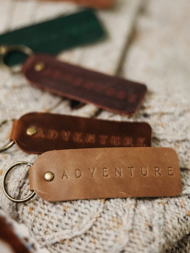 encouraging leather keyring. brown keyrings laid atop a blanket, embossed with the word 'adventure'.