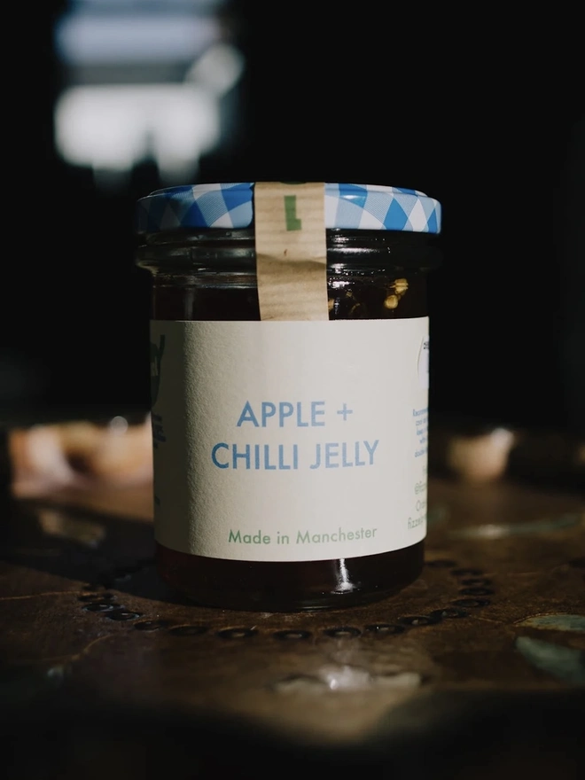 Jar Of Apple & Chilli Jelly, jar of jelly with a blue lid on a decorated plate on a table. 