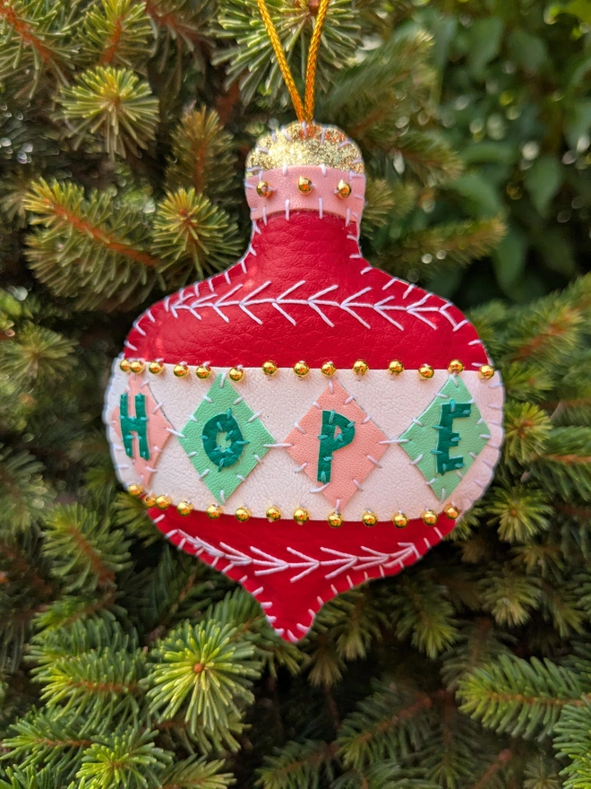 Hand-stitched red, pink and green vegan leatherette Christmas bauble decoration featuring the word ‘HOPE’, white embroidered details, gold bead embellishments and a gold hanging loop, displayed on a Christmas tree.