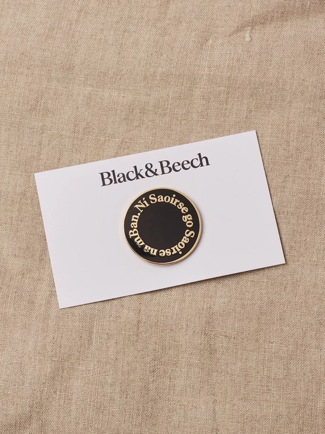A black round enamel pin with the words Ní Saoirse go Saoirse na mBan written around the edge in gold on white backing card 