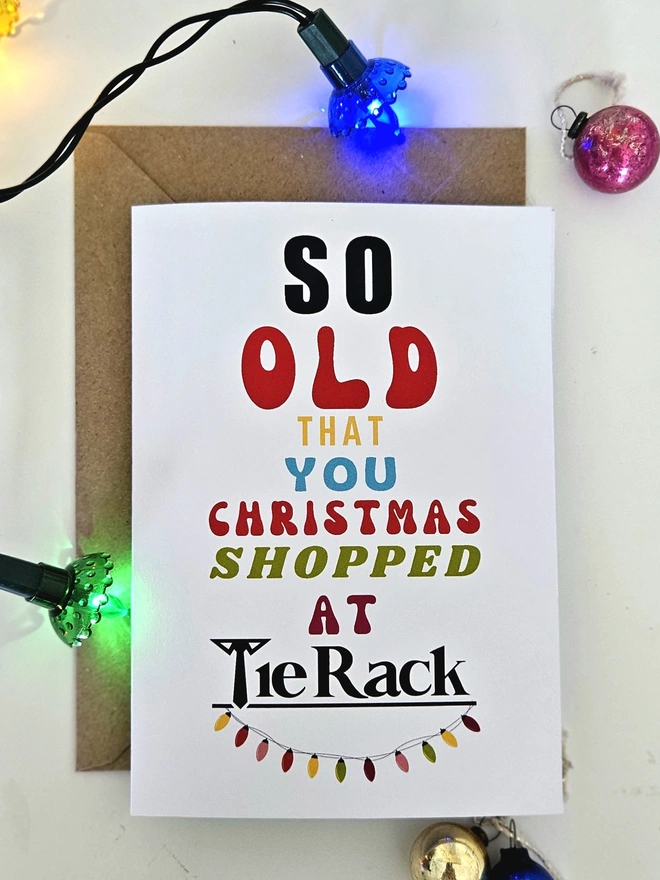 A photograph of the front of a humorous A6 greeting card illustrated using brightly coloured text and the retro shop logo. The text says so old  that you Christmas shopped at Tie Rack with string lights