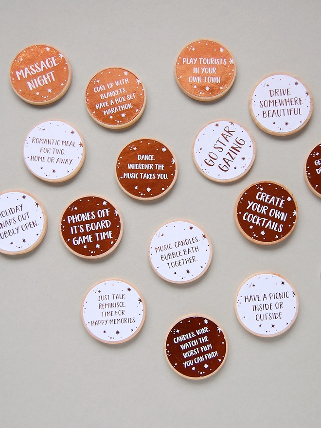 15 wooden tokens each with a copper foiled label that have a date night idea printed on lay on a grey background.
