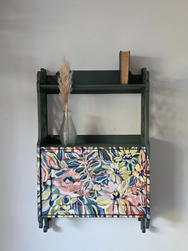 Vintage wall cabinet painted earthy matt green pictured against a white wall. The front has been handpainted with florals and stems in tan, ochre, navy and shades of peach