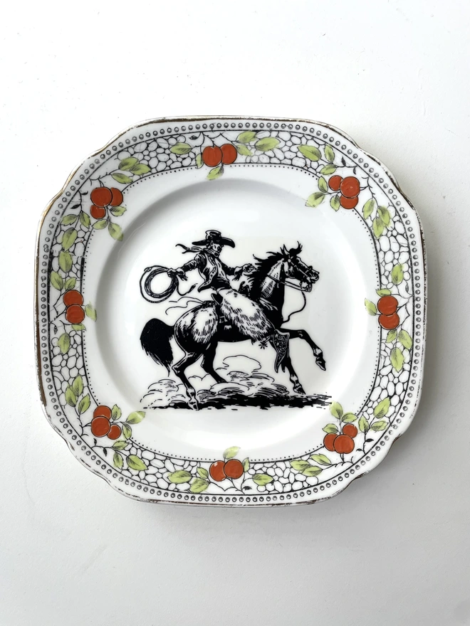vintage plate hanging on a white wall, it has a decorative border, with a printed vintage illustration of a cowboy in the middle 