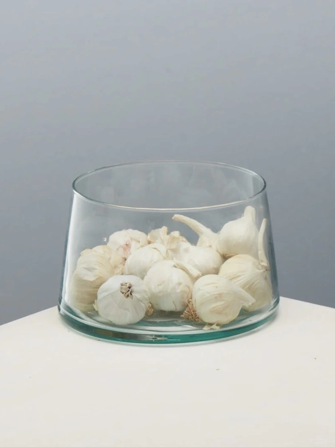 Tall Glass Dish, a tall glass dish sitting on a white surface against a baby blue backdrop. It is filled by garlic. 