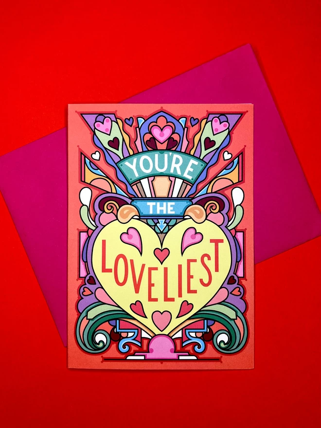 A greetings card with the phrase “You’re the Loveliest” surrounded by an abstract multi-coloured design and lots of hearts, sits on top of a pink envelope on a red backdrop.