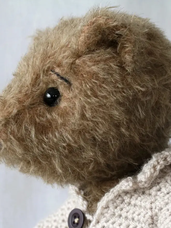 Brown teddy bear sitting upright, wearing a cream knitted cardigan with three buttons, against a plain light background.