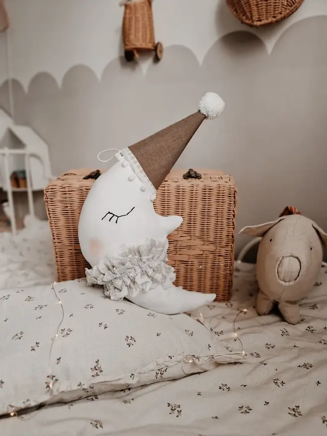 Hanging Moon Pillow Decoration, a hanging moon pillow decoration sitting against a wooden hamper accompanied by other bedding accessories. 