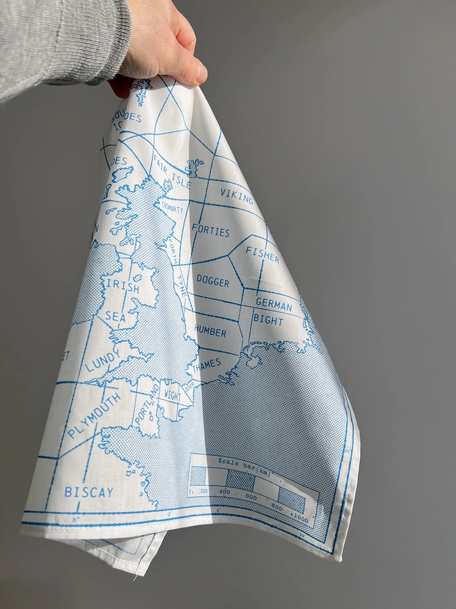 A white hand holds a blue printed Mr.PS Shipping Map Handkerchief against a sunlit wall