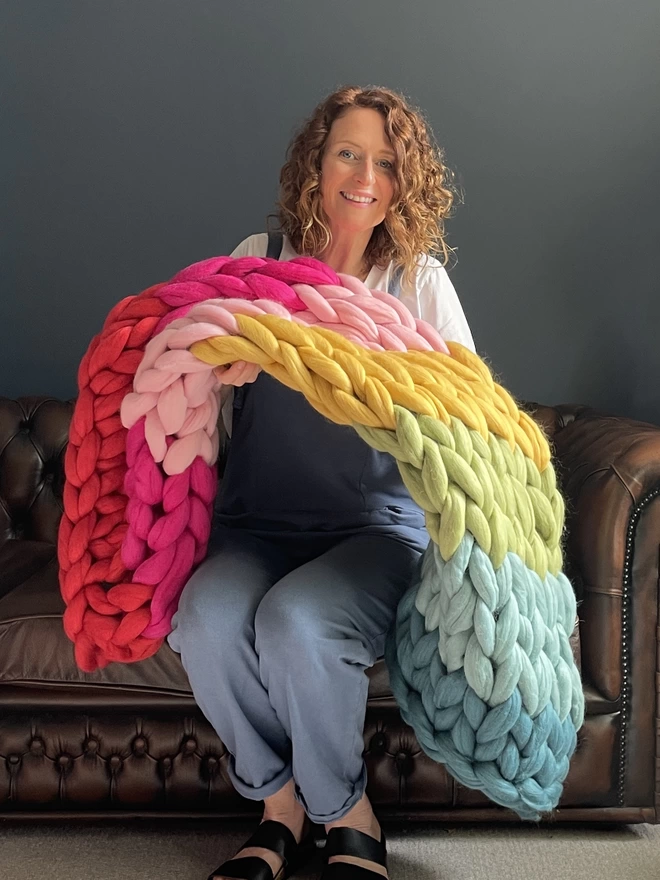 Mizz, a white woman in her forties, is smiling at the camera, she is sitting on a brown leather sofa holding a rainbow coloured merino giant knitted blanket up in from of her