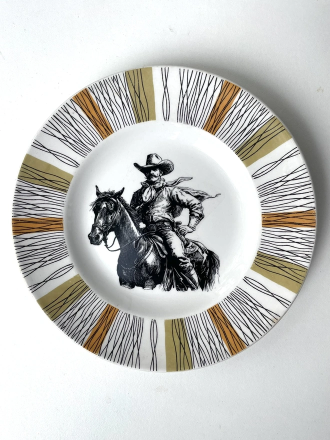 vintage plate hanging on a white wall, it has a decorative border of grey and blue leaves, with a printed vintage illustration of a cowboy in the middle 