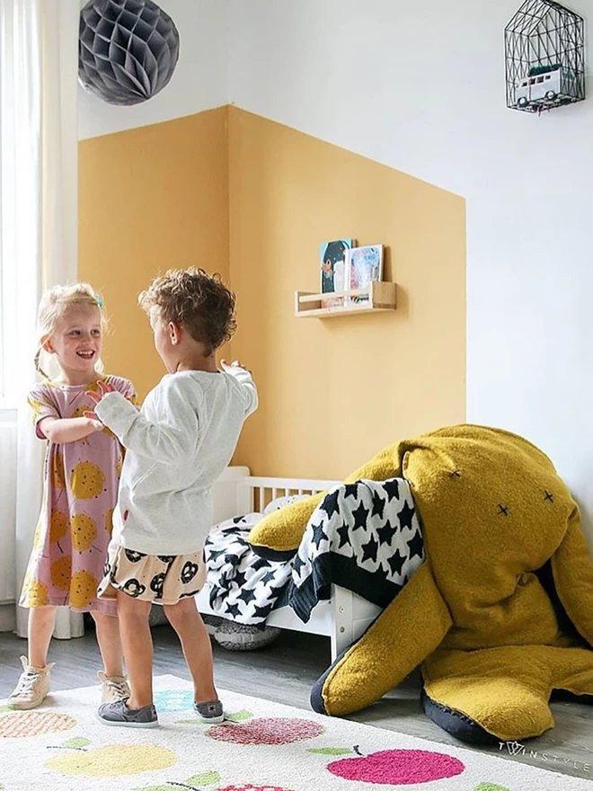 Two children are laughing and playing on the left of the frame. On the right is a childrens bed with ablack and white star blanket spread over it. At the foot of the bed sits a giant mustard yellow bunny rabbit.