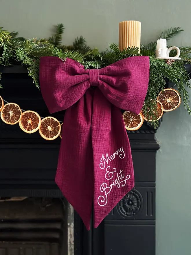 A handmade Christmas bow made from deep burgundy cotton with the words "Merry & Bright" on one tail is hung from a black mantlepiece that is decorated for Christmas with greenery, orange slice garland, and candles.