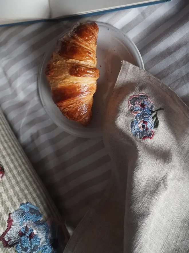 Natural linen napkin embroidered with blue and pink flowers, styled for breakfast with coordinating embroidered homeware.
