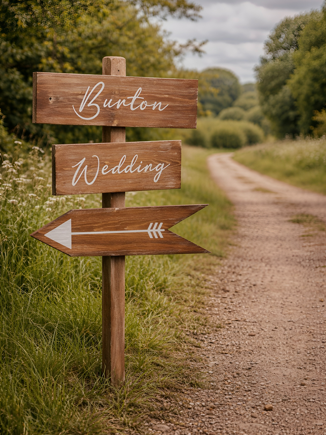 a rustic wedding direction sign with an arrow pointing to the left on a countryside path leading to a wedding venue