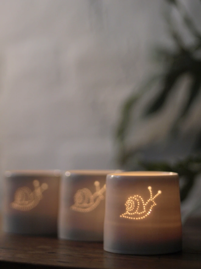 Group of porcelain tealight holders, all with small holes pierced in the clay, allowing the light to shine through the hole and form an image. The dots are in the pattern of a snail facing right, showing the curly shell.