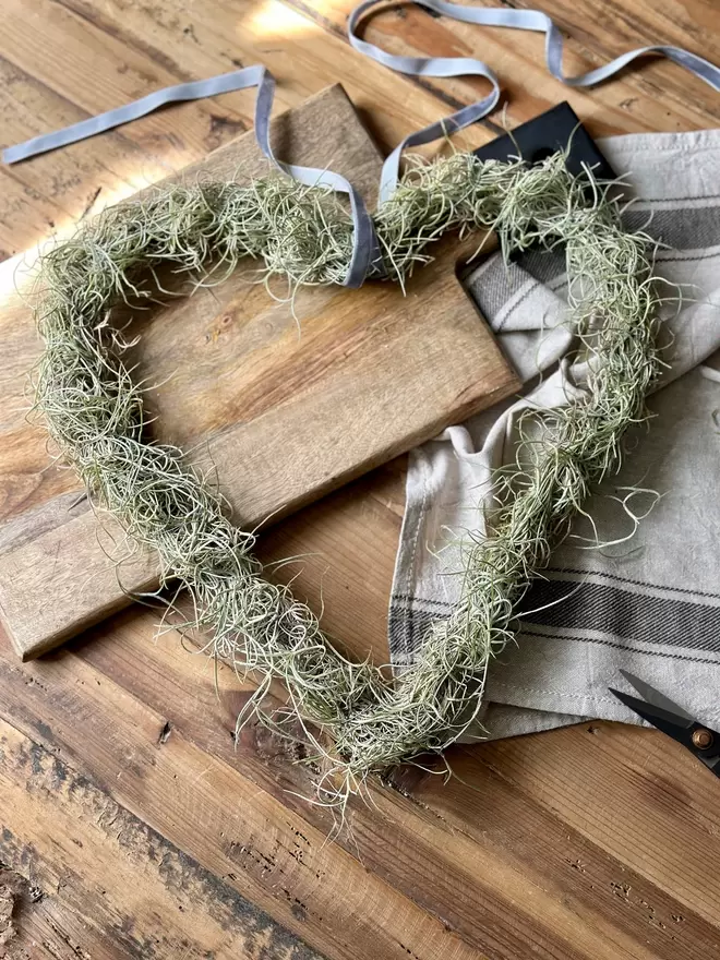 A wire Love Heart arrangement made of Tillandsia, finished  at the top with a thin ribbon for hanging, it lays across a wooden board, a ruffled vintage tea towel sits in the background along with a pair of small scissors.