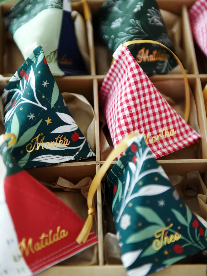A Christmas decorations box full of handmade fabric fortune cookie decorations, each from a different festive fabric and with a personalised name in gold lettering, with a gold ribbon. 