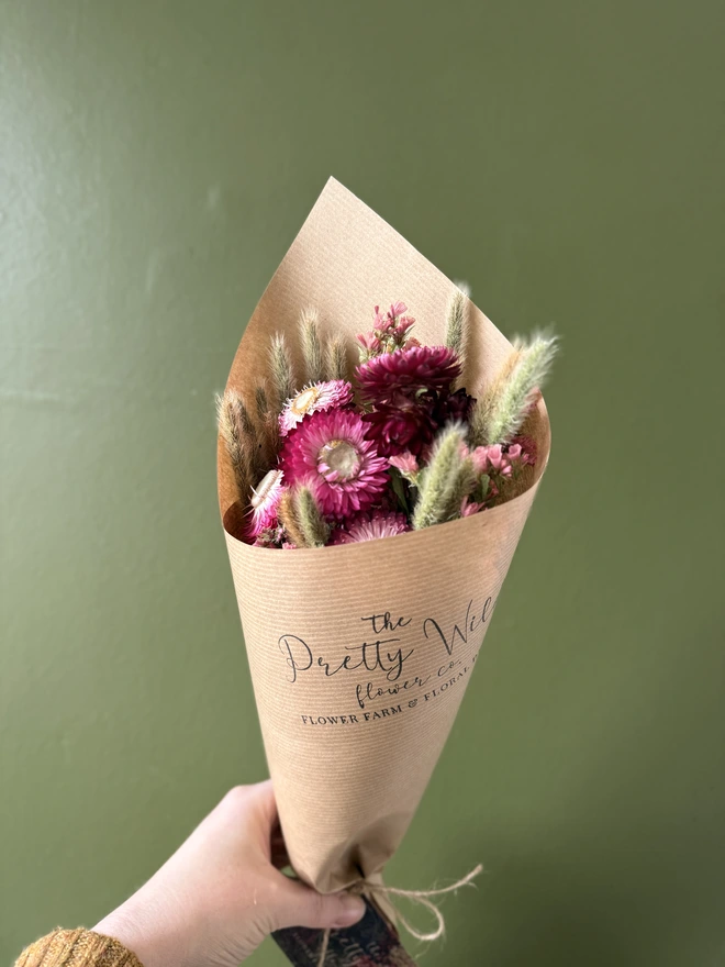 a bunch of dried flowers in bright colours wrapped in kraft paper held by a person in a mustard coloured jumper with their left arm against a green wall