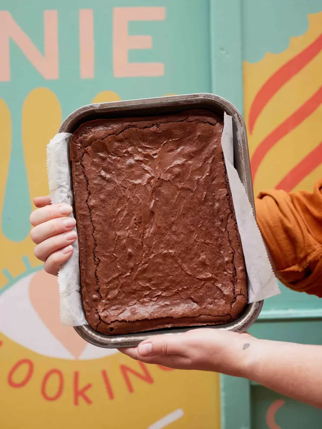 Founder Hetty holding a freshly baked gluten free classic fudge brownies in a baking tray against a colourful background