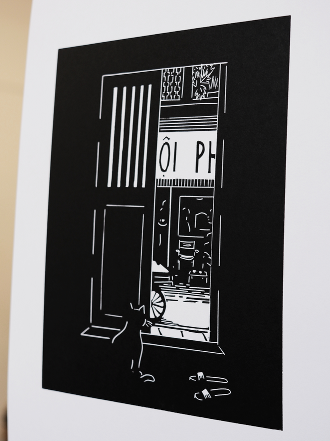 A black and white print depicting a cat watching a street in Vietnam from behind a shutter door. Slippers are by the door and a coffee shop across the road. Done in a simplistic and stylistic way