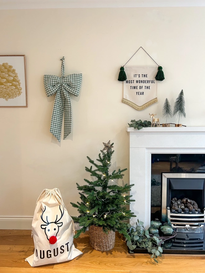 Christjmas Present Sack with Reindeer design and red pom pom nose sitting next to a Christmas Tree and fireplace