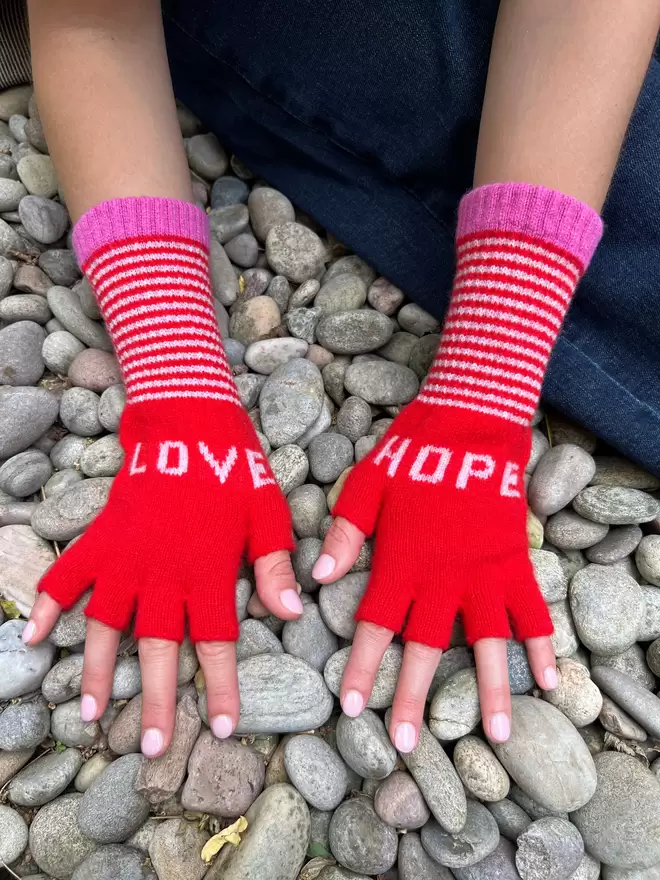 A classic red fingerless knitted glove with the words LOVE and HOPE in dusky pink and stripe cuff with a bubblegum pink edge detail. Made from 100% Lambswool with Wool spun in Yorkshire. 