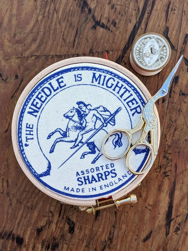 Royal blue coloured 'the needle is mightier' embroidery hoop needle book depicting a soldier on horse back with a needle as a sword shown with stork scissors and cotton reel
