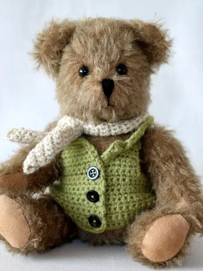 Handmade brown teddy bear with a white scarf wearing a green cardigan, sitting against a plain background.