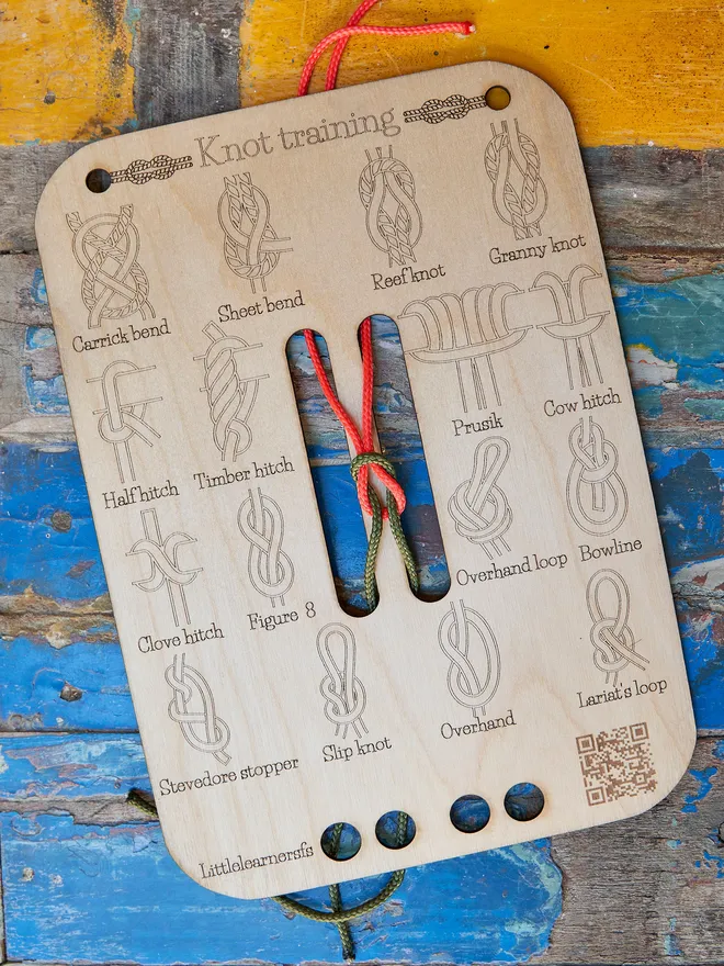 Wooden knot training board with engraved knot diagrams and red and black ropes for practice, displayed outdoors