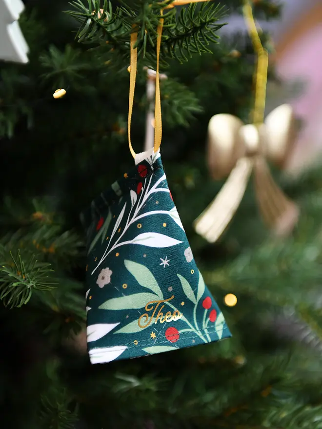 A handmade fabric fortune cookie Christmas decoration in green festive foliage print, personalised with the name “Theo” in gold, hangs from a golden ribbon on a Christmas tree branch.