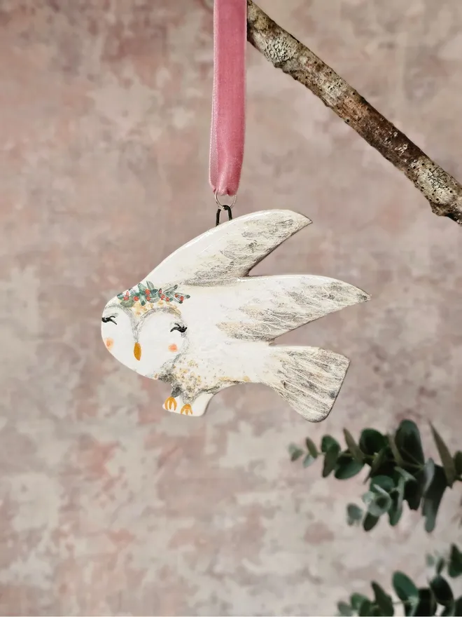 Decorative owl ornament with floral crown hanging from a branch on a soft pink background.