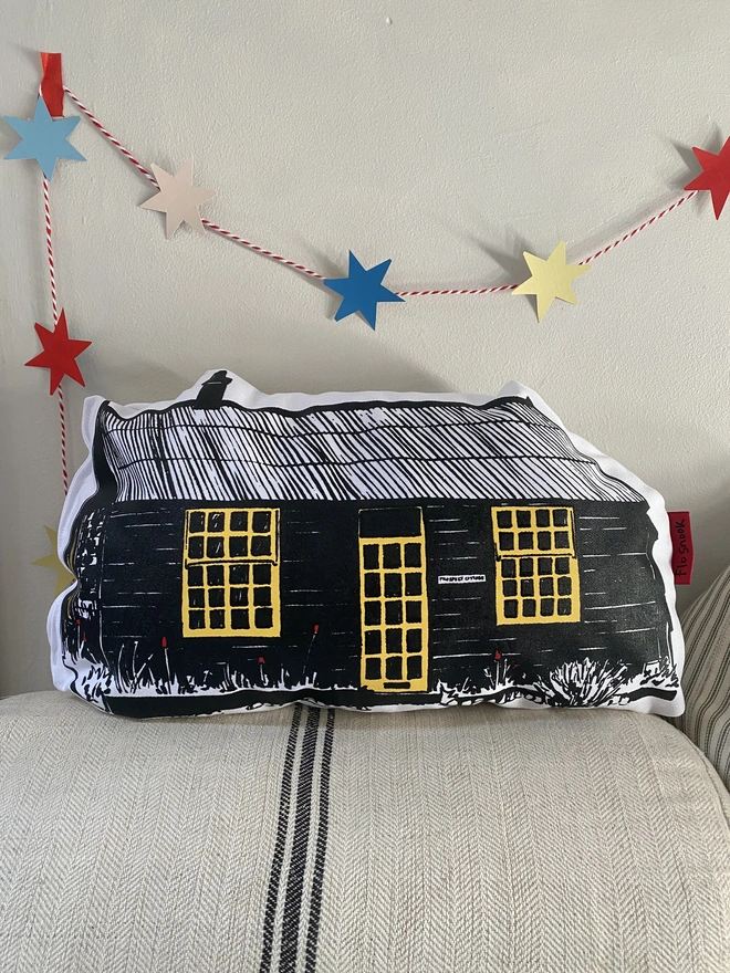 A prospect cottage handmade cushion by artist Flo Snook. A black and yellow cushion in the shape of prospect cottage, sitting on a linen cushion with a stars garland hanging behind