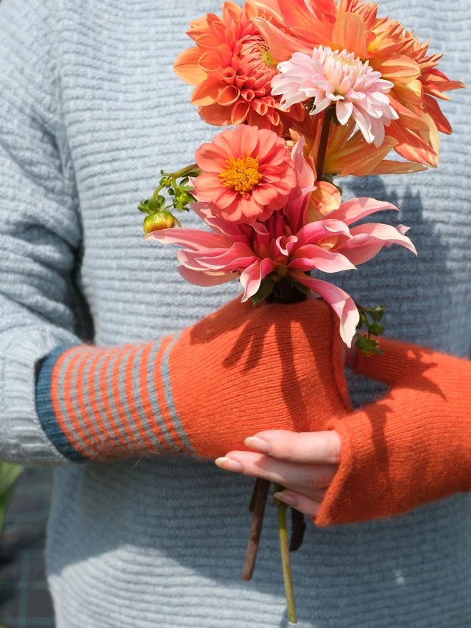 JAFFA ORANGE FINGERLESS MITTEN WITH A TEAL COLOUR STRIPE CUFF SUPER SOFT AND WARM WHILE LEAVING HANDS FREE ON THIS IMAGE HANDS ARE HOLDING A BEAUTIFUL SUNNY POSY OF PINK AND ORANGE DAHLIAS