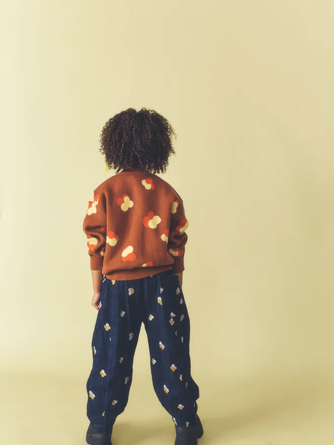 Back view of a child against a pale yellow backdrop, showing the reverse of a Studio Koter chestnut floral intarsia cardigan and embroidered dark denim trousers with a rear patch pocket.