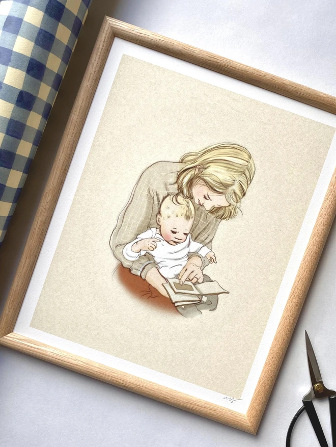 a photograph of a framed mummy and me art print, an oak frame laid flat on wrapping paper, the print is a hand drawn illustration of a mother with blond hair with a bay on her lap looking at a baby book