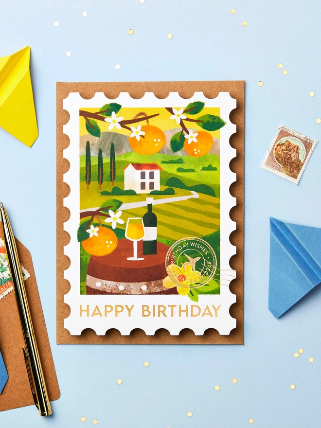 Stamp-shaped greeting card featuring an illustration of Tuscany. The card is on a blue background with paper aeroplanes and stationery around it.