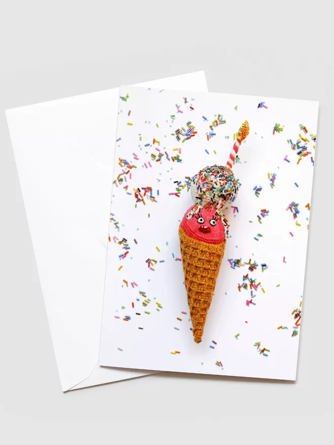 Image of Kates Birthday Cone Card seen with its white envelope. On the front of the card is a waffle cone with pink and white ice cream covered in sprinkles and a stripey candle.