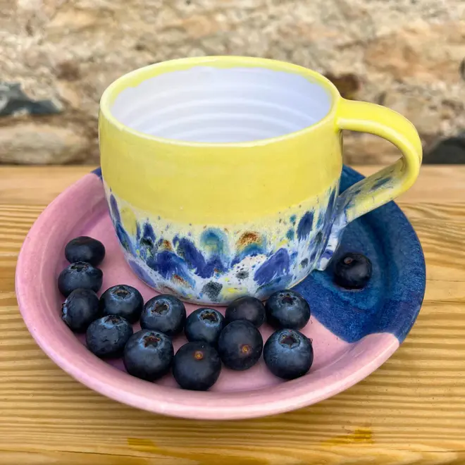 Shoreline Large Round Mug, a colourful mug on a wooden surface against a brick backdrop. It is accompanied by fruits. 