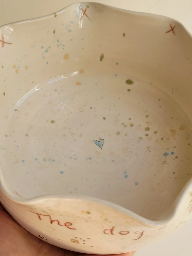 ivory ceramic dog bowl with 'the dog's dinner' written on the side with brown green and blue splatters and pawprints held in a hand with a blue heart in the centre of the bowl