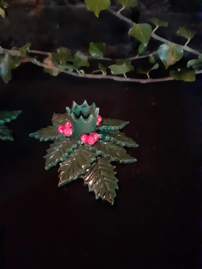 Angled view of a holly candle holder showing overlapping green leaves, red berries and the pointed rim of the candle cup.