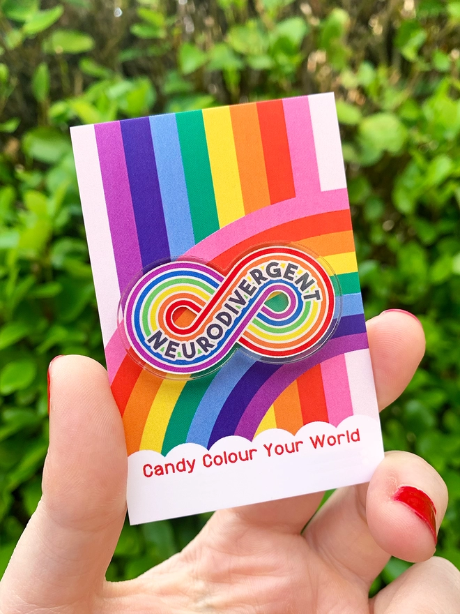 image shows a hand holding a rainbow business card. pinned to the card is an acrylic badge in the shape of a rainbow stripe infinity loop. printed across the loop is the word 'Neurodivergent'