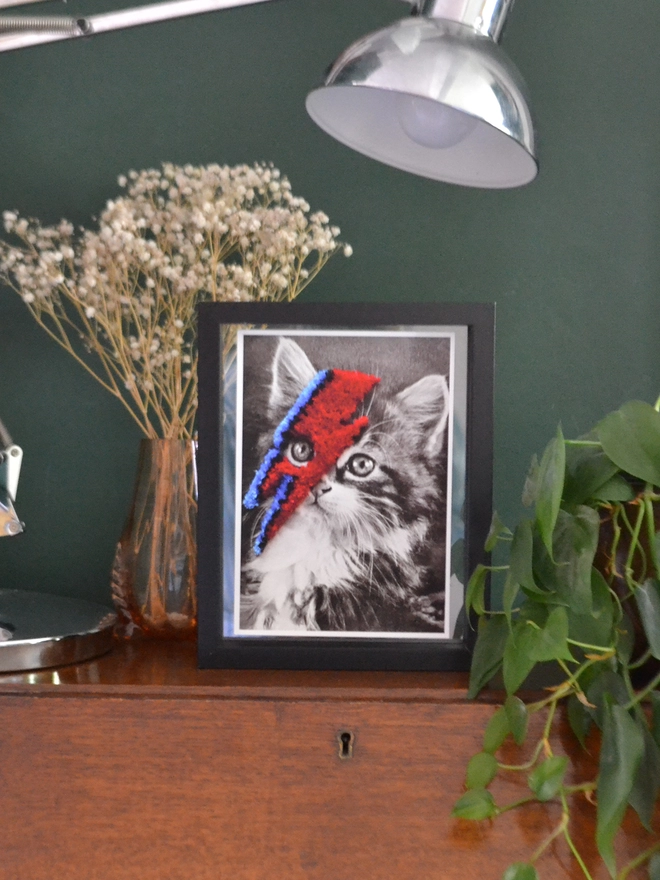 Black and white cat print with red and blue embroidered bolt in frame on desk