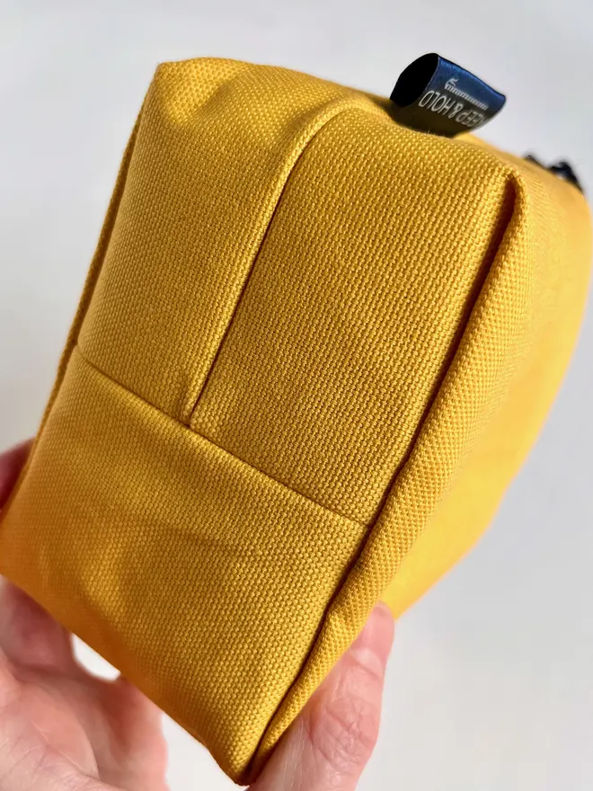 Hand holding up a small yellow fabric bag to show the rectangular, boxed bottom construction.