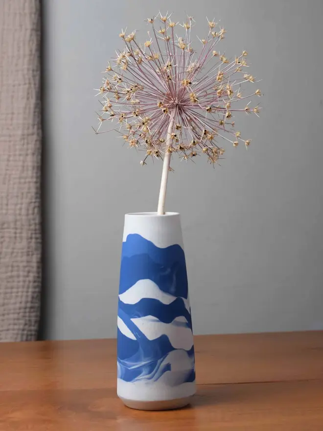 vase on wooden table, vase has streaky pattern in shades of blue and holds dry plant