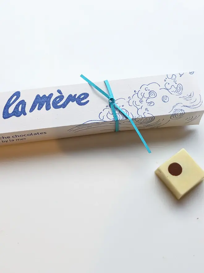 marc de champagne ganache chocolates. a white rectangular box with a blue illustrated wave, photographed next to a square white ganache chocolate.