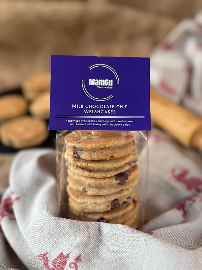 Chocolate Chip Welsh Cakes in clear packaging with a purple tag on a busy kitchen work top includung welsh dragon table cloth and wooden rolling pin