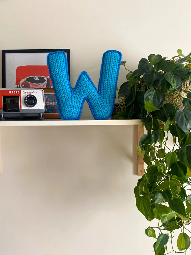 Crochet cushion shaped like the letter W standing on a shelf, in Light Blue and Blue